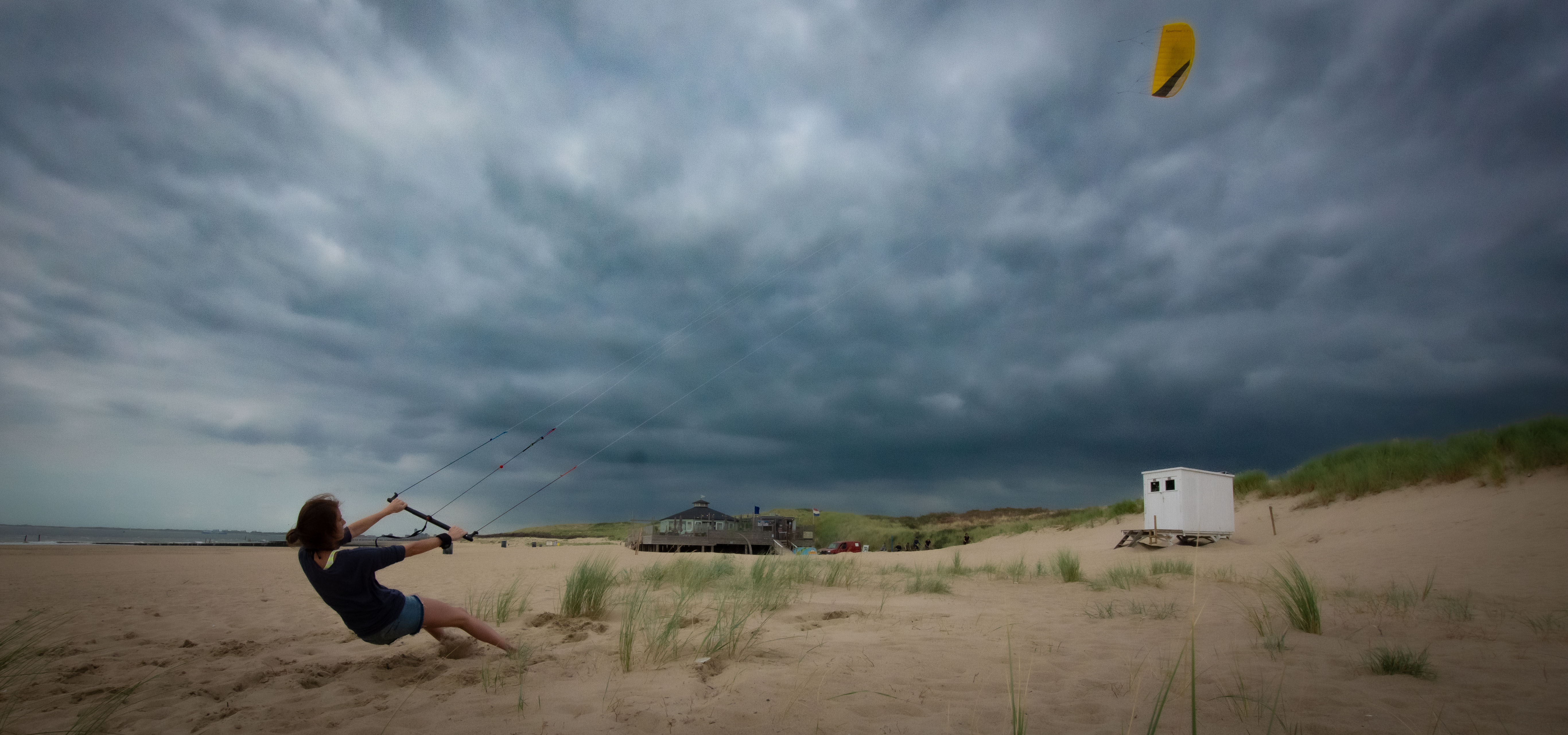 ein Strandpanoroma mit dramatischem Wolkenhimmel, vor dem eine junge Frau einen Kite steuert